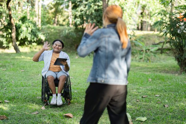 A young man in a wheelchair waves to a friend in a sunny park, highlighting friendship and inclusivity.