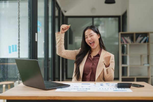 Young asian businesswoman celebrating success with raised arms in her office, radiating joy and excitement after reaching her goals
