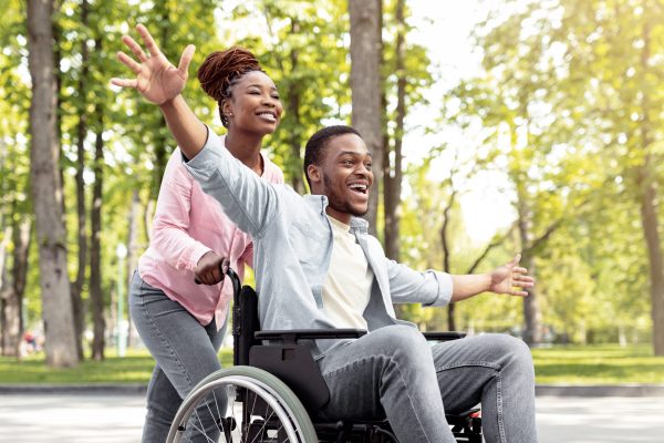 Happy black disabled guy in wheelchair on walk with his loving girlfriend outdoors, having fun, spending time together. African American impaired couple enjoying each other's company at green park