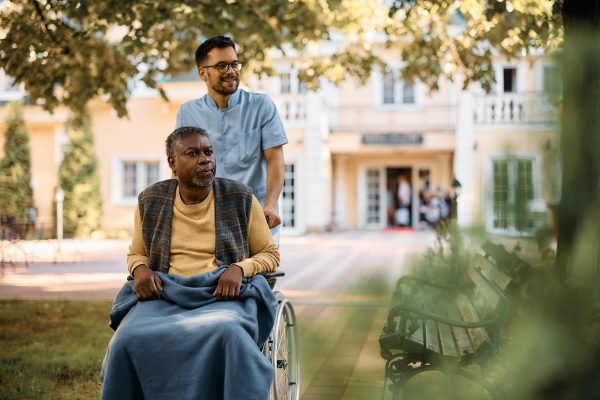 Young healthcare worker pushing senior black man in wheelchair through nursing home's park.